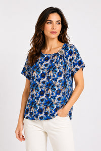 Εμπριμέ μπλούζα με μπλε floral print, κοντό μανίκι, στρογγυλή λαιμόκοψη και διακοσμητική καρφίτσα στον ώμο.