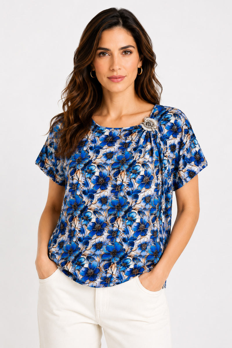 Εμπριμέ μπλούζα με μπλε floral print, κοντό μανίκι, στρογγυλή λαιμόκοψη και διακοσμητική καρφίτσα στον ώμο.