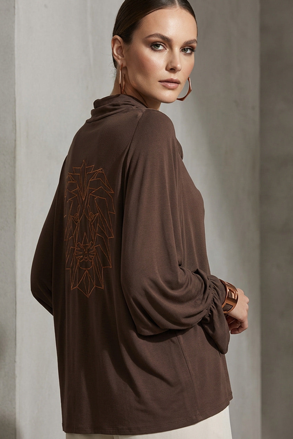 Καφέ μπλούζα ντραπέ με μακριά μανίκια και διακοσμητικό τύπωμα λιονταριού στην πλάτη, φορεμένη σε μοντέλο.