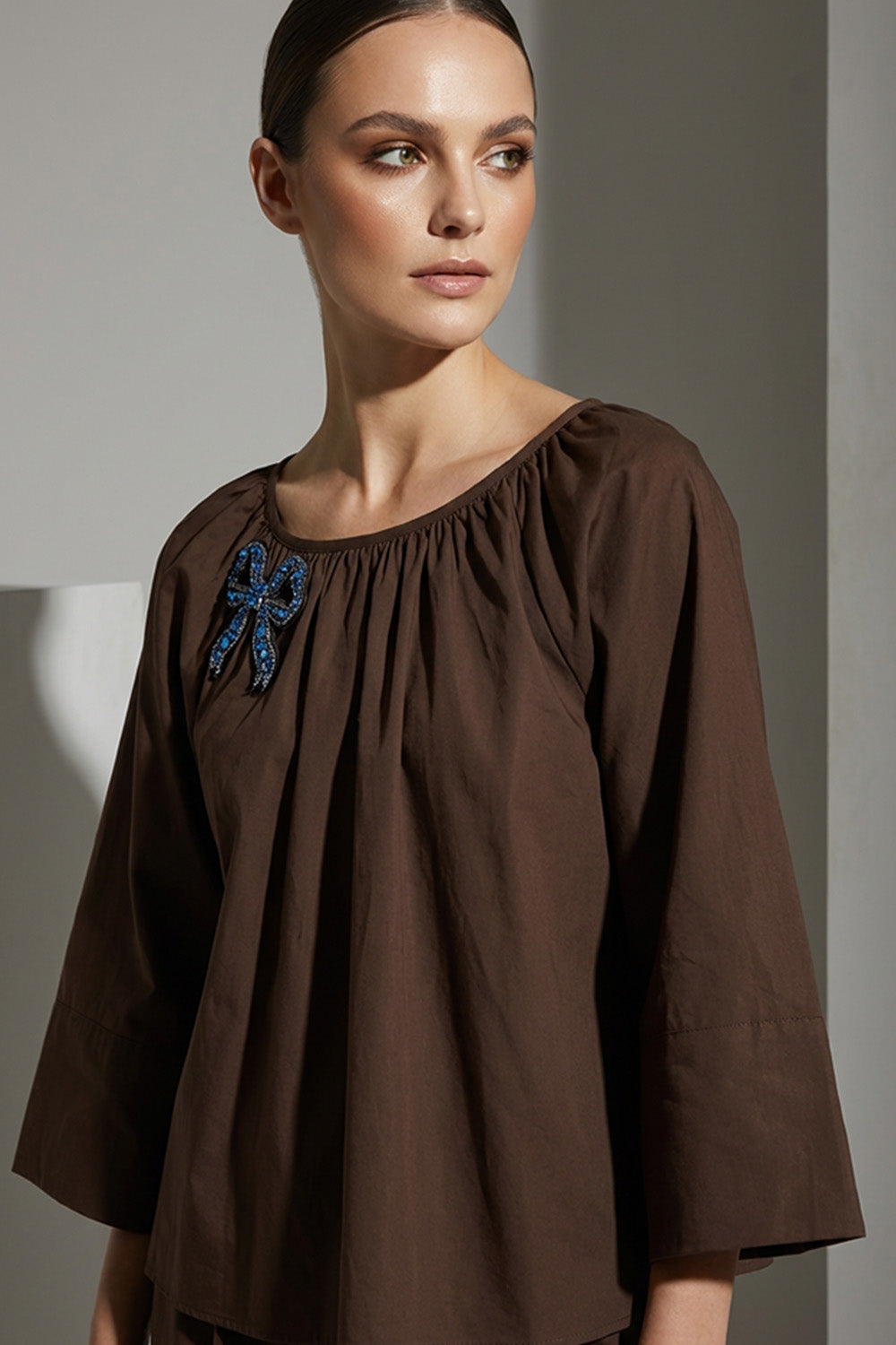 Καφέ γυναικεία μπλούζα με στρογγυλή λαιμόκοψη και κεντημένο μπλε φιόγκο στο στήθος.