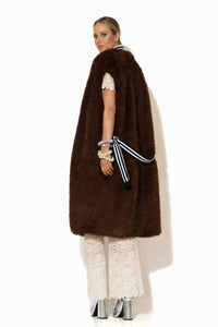Μακρύ αμάνικο παλτό από συνθετική γούνα σε σκούρο καφέ χρώμα, με πλούσια υφή και ίσια γραμμή, φορεμένο σε κομψό σύνολο.