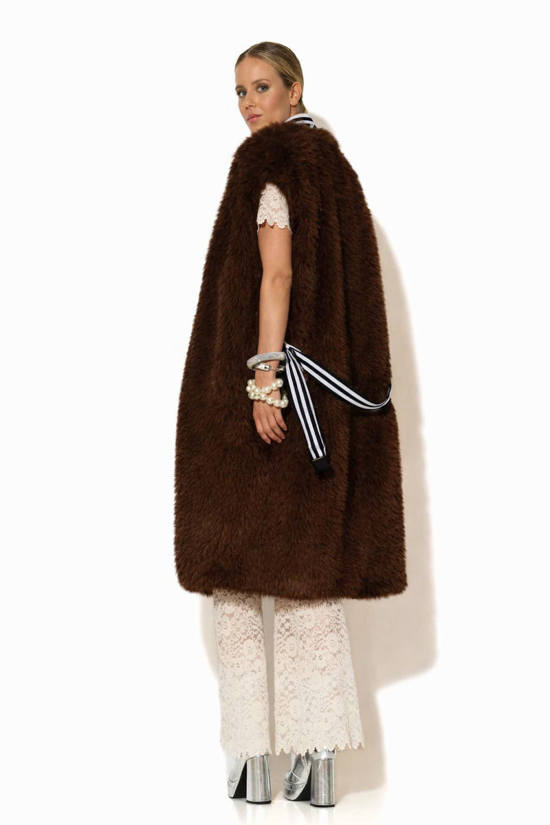 Μακρύ αμάνικο παλτό από συνθετική γούνα σε σκούρο καφέ χρώμα, με πλούσια υφή και ίσια γραμμή, φορεμένο σε κομψό σύνολο.