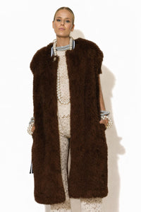 Μακρύ αμάνικο παλτό από συνθετική γούνα σε σκούρο καφέ χρώμα, με πλούσια υφή και ίσια γραμμή, φορεμένο σε κομψό σύνολο.