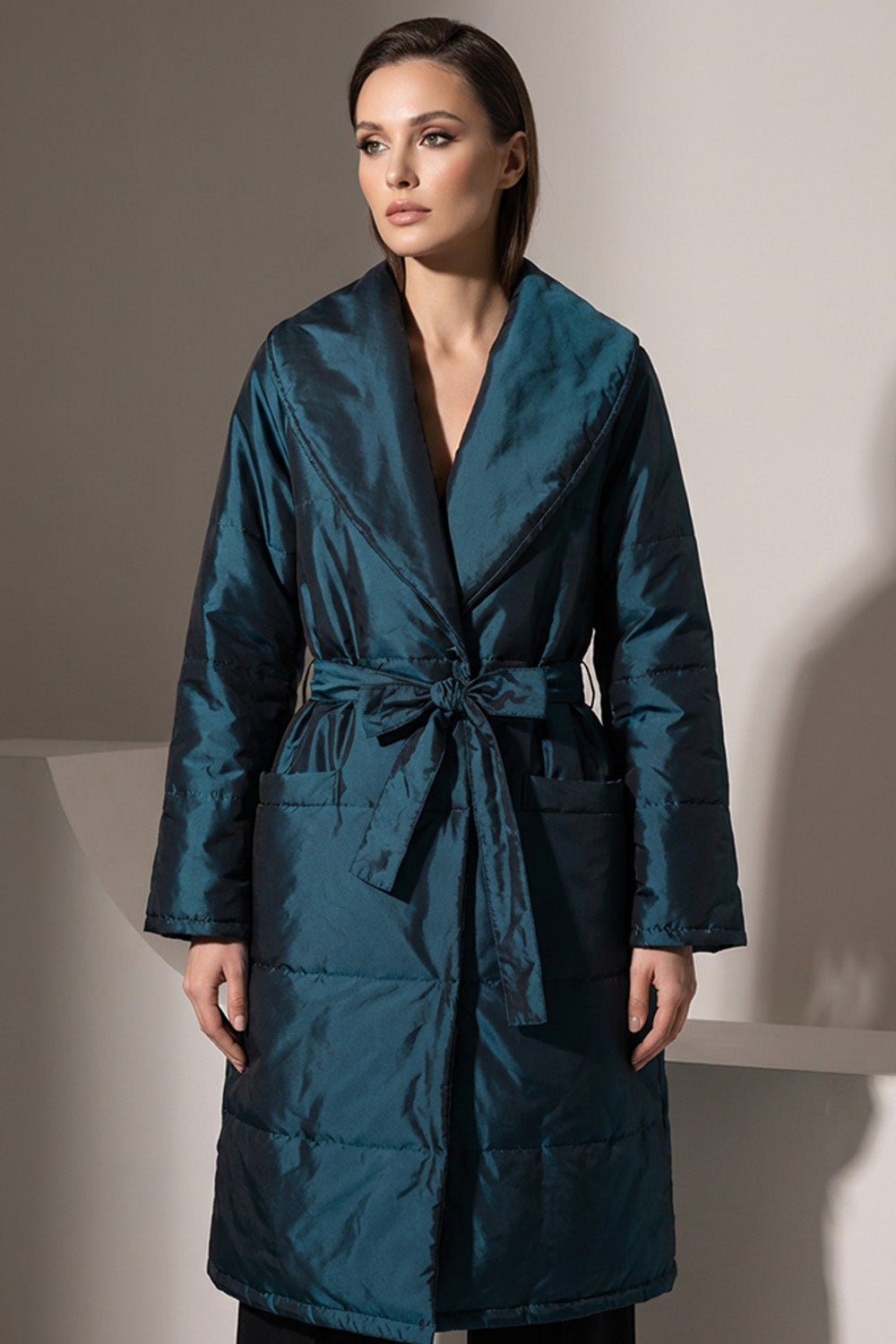 Γυναικείο μακρύ καπιτονέ παλτό σε πετρόλ χρώμα με ζώνη στη μέση και φαρδύ γιακά.