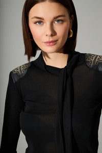 Μαύρη γυναικεία μπλούζα με μακριά μανίκια, χρυσές λεπτομέρειες στους ώμους και δέσιμο στον λαιμό, φορεμένη σε μοντέλο σε studio φωτογράφιση