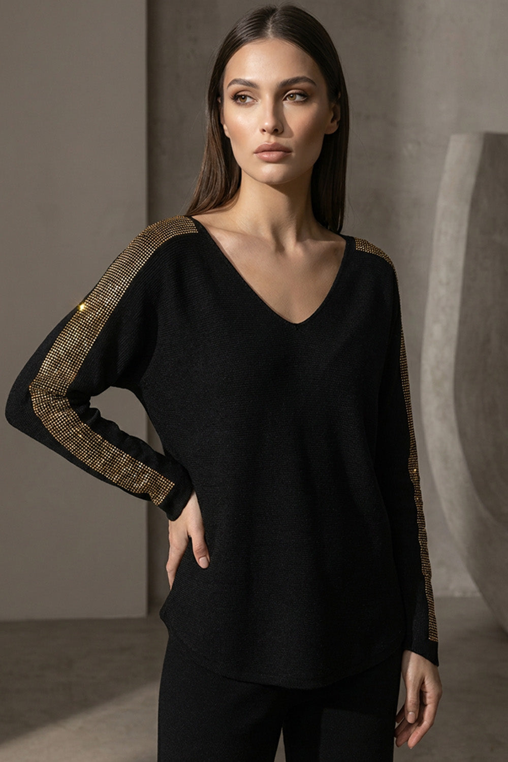 Μαύρη πλεκτή μπλούζα με V λαιμόκοψη και χρυσές λεπτομέρειες στους ώμους και μανίκια, φορεμένη από μοντέλο studio.
