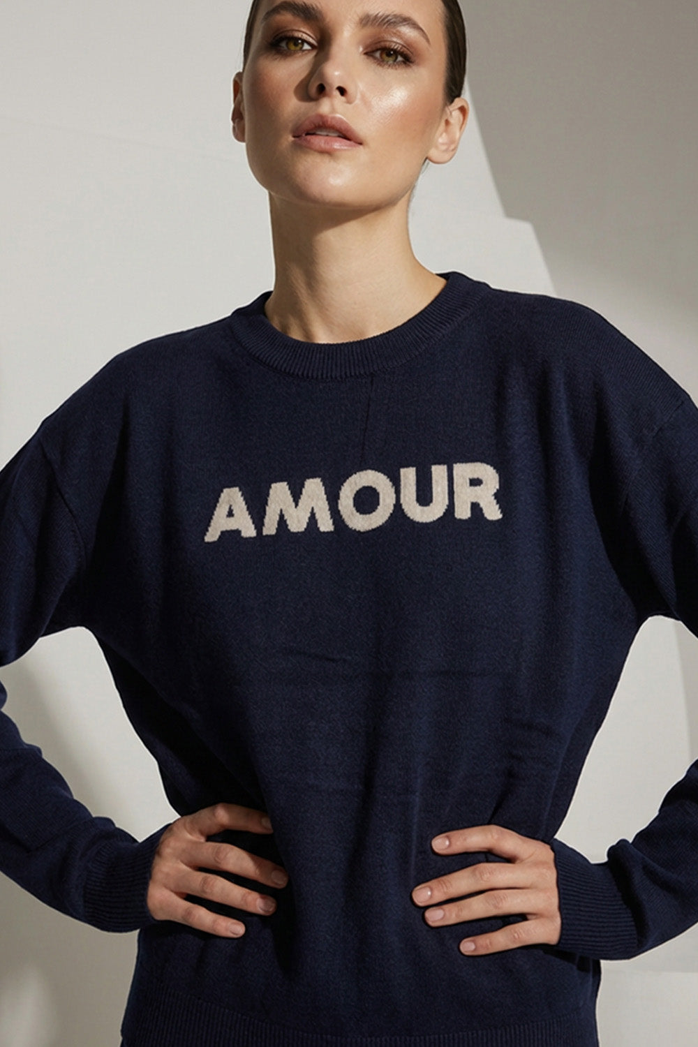 Μπλε πλεκτή μπλούζα με επιγραφή Amour στο μπροστινό μέρος, φορεμένη από μοντέλο σε minimal studio φόντο.