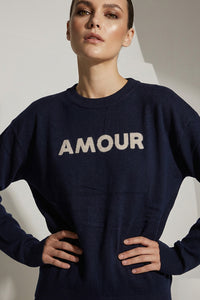Μπλε πλεκτή μπλούζα με επιγραφή Amour στο μπροστινό μέρος, φορεμένη από μοντέλο σε minimal studio φόντο.