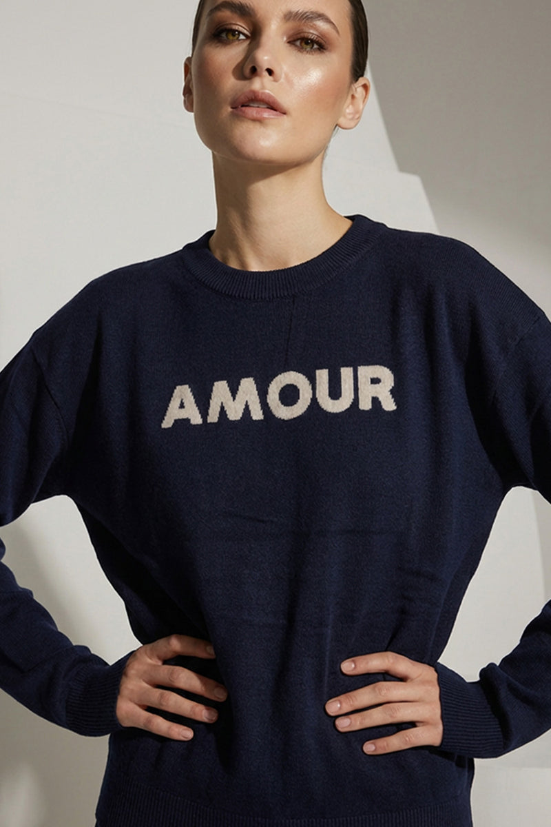 Μπλε πλεκτή μπλούζα με επιγραφή Amour στο μπροστινό μέρος, φορεμένη από μοντέλο σε minimal studio φόντο.