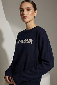 Μπλε πλεκτή μπλούζα με επιγραφή Amour στο μπροστινό μέρος, φορεμένη από μοντέλο σε minimal studio φόντο.