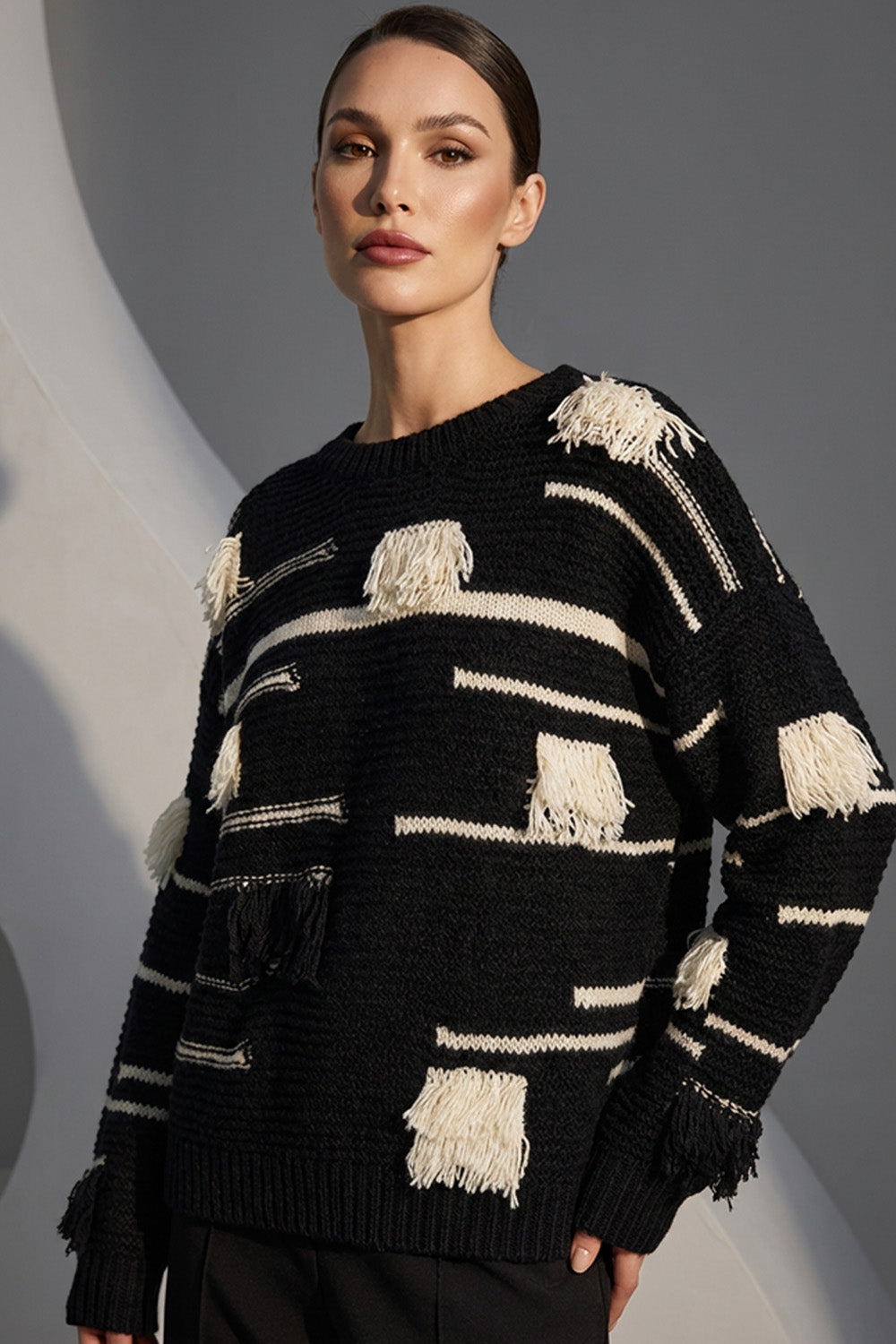 Μαύρη πλεκτή μπλούζα με εκρού ρίγες και διακοσμητικά κρόσσια, φορεμένη από μοντέλο σε studio φόντο.