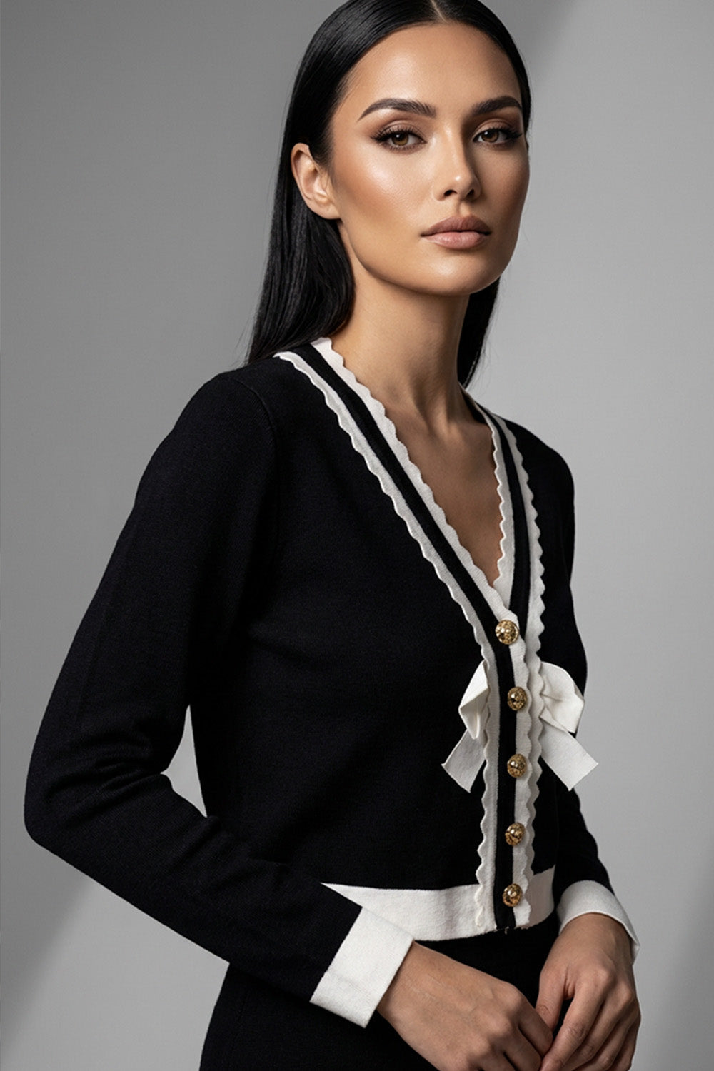 Μαύρη πλεκτή ζακέτα με λευκές λεπτομέρειες, χρυσά κουμπιά και διακοσμητικούς φιόγκους μπροστά.
