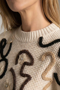 Πλεκτή εκρού μπλούζα με ανάγλυφο κεντημένο σχέδιο σε μαύρο και καφέ, στρογγυλή λαιμόκοψη και μακριά μανίκια, φορεμένη από μοντέλο σε studio φωτογράφιση.