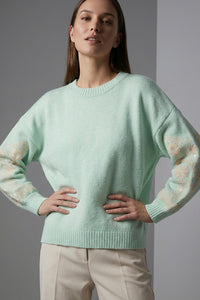 Πλεκτή mint μπλούζα με στρογγυλή λαιμόκοψη, μακρύ μανίκι με κεντημένο floral μοτίβο και άνετη γραμμή, φορεμένη με ανοιχτόχρωμο παντελόνι.