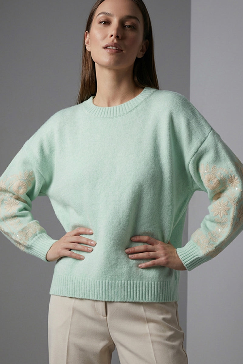 Πλεκτή mint μπλούζα με στρογγυλή λαιμόκοψη, μακρύ μανίκι με κεντημένο floral μοτίβο και άνετη γραμμή, φορεμένη με ανοιχτόχρωμο παντελόνι.