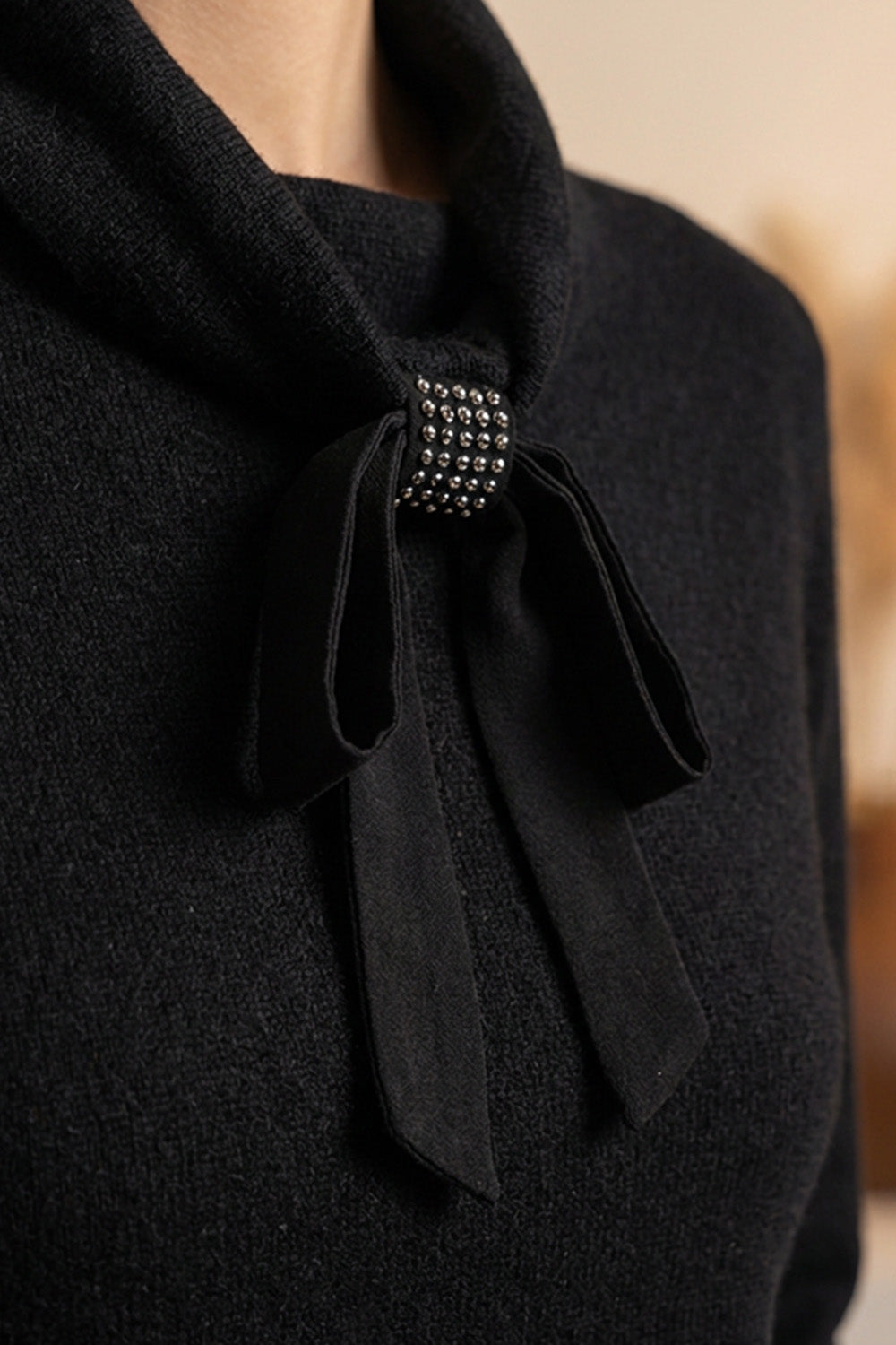 Μαύρη πλεκτή μπλούζα με φιόγκο στον λαιμό και μεταλλική διακοσμητική λεπτομέρεια, φορεμένη από μοντέλο σε minimal studio σκηνικό.