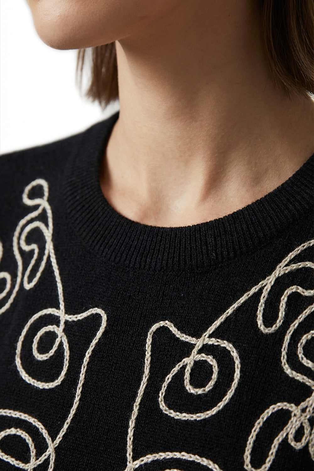 Μαύρη πλεκτή μπλούζα με εκρού κεντημένο καλλιτεχνικό σχέδιο στο μπροστινό μέρος, φορεμένη από μοντέλο σε minimal studio περιβάλλον.
