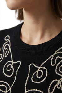 Μαύρη πλεκτή μπλούζα με εκρού κεντημένο καλλιτεχνικό σχέδιο στο μπροστινό μέρος, φορεμένη από μοντέλο σε minimal studio περιβάλλον.