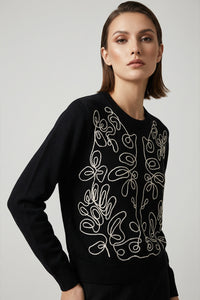 Μαύρη πλεκτή μπλούζα με εκρού κεντημένο καλλιτεχνικό σχέδιο στο μπροστινό μέρος, φορεμένη από μοντέλο σε minimal studio περιβάλλον.