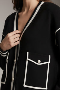 Μαύρη πλεκτή ζακέτα με λευκά τελειώματα, V λαιμόκοψη και διακοσμητικές τσέπες, φορεμένη από μοντέλο.