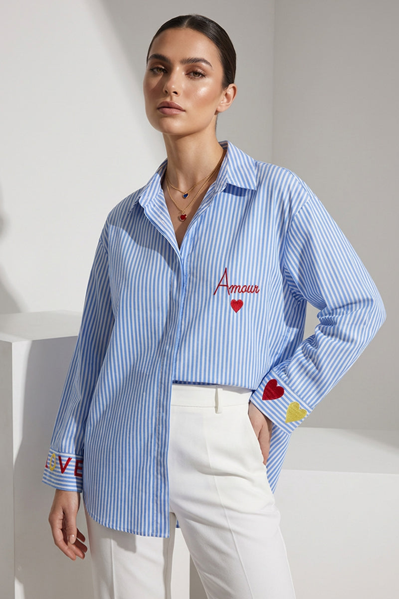 Γυναικείο ριγέ γαλάζιο-λευκό πουκάμισο με κεντημένο μήνυμα και λεπτομέρειες στα μανίκια, φορεμένο με λευκό παντελόνι.