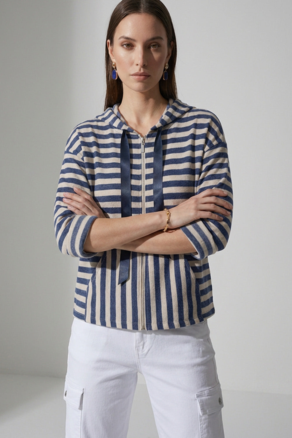Γυναικεία ριγέ ζακέτα με κουκούλα και φερμουάρ σε μπλε και εκρού αποχρώσεις, φορεμένη με λευκό cargo παντελόνι.