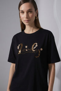 Γυναικείο μαύρο t-shirt με κεντημένο λογότυπο και διακοσμητικές χάντρες, φορεμένο με λευκό παντελόνι.