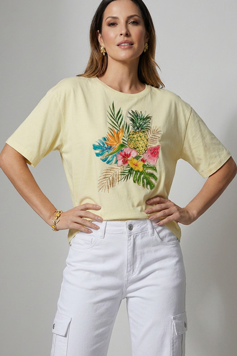 Γυναικείο κίτρινο t-shirt με τροπικό τύπωμα φύλλων και φρούτων, ελαστικό ύφασμα και κοντό μανίκι, φορεμένο με λευκό cargo παντελόνι.