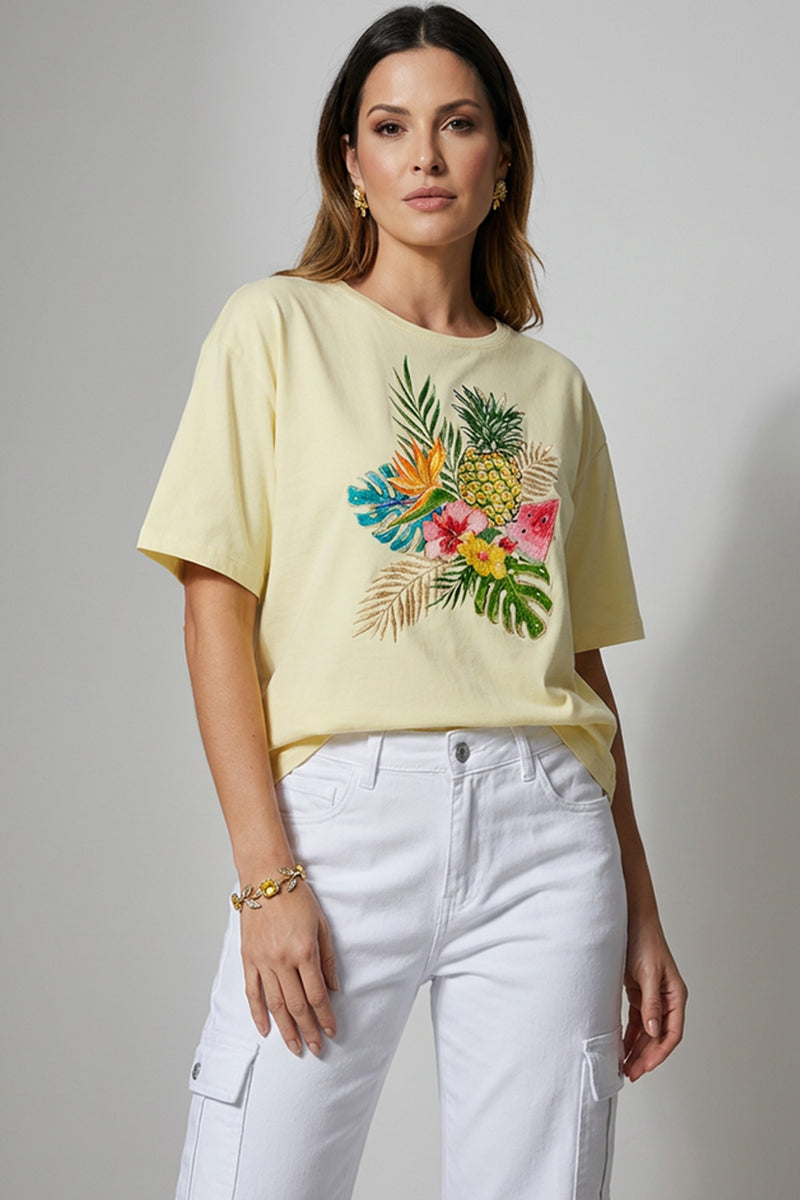 Γυναικείο κίτρινο t-shirt με τροπικό τύπωμα φύλλων και φρούτων, ελαστικό ύφασμα και κοντό μανίκι, φορεμένο με λευκό cargo παντελόνι.