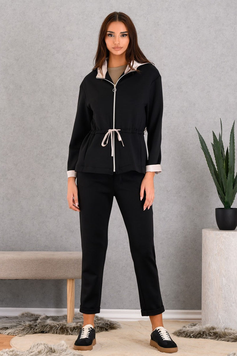 Woman wearing a black outfit with a white shirt underneath, standing in a room with a gray wall and a plant.